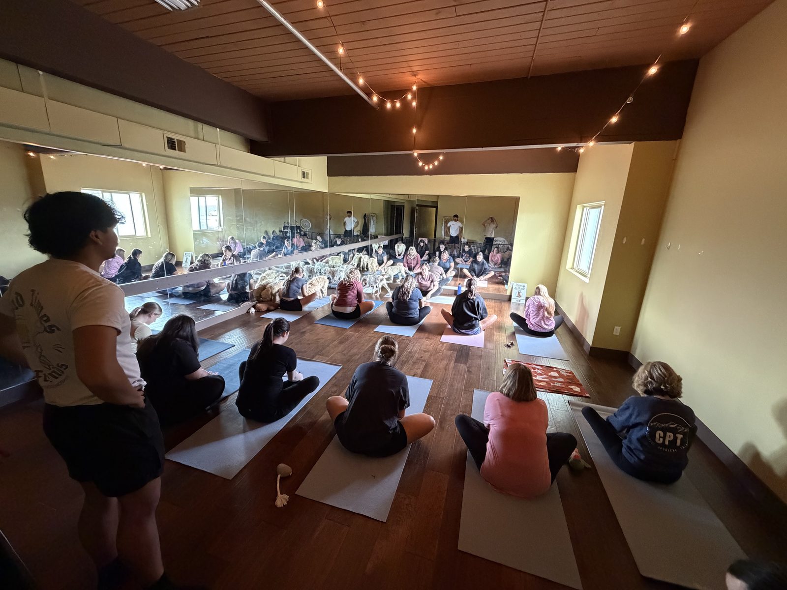 A real Paws & Pose class in the upstairs Prana Salon studio — guests on mats facing the mirrored wall, string lights warm overhead
